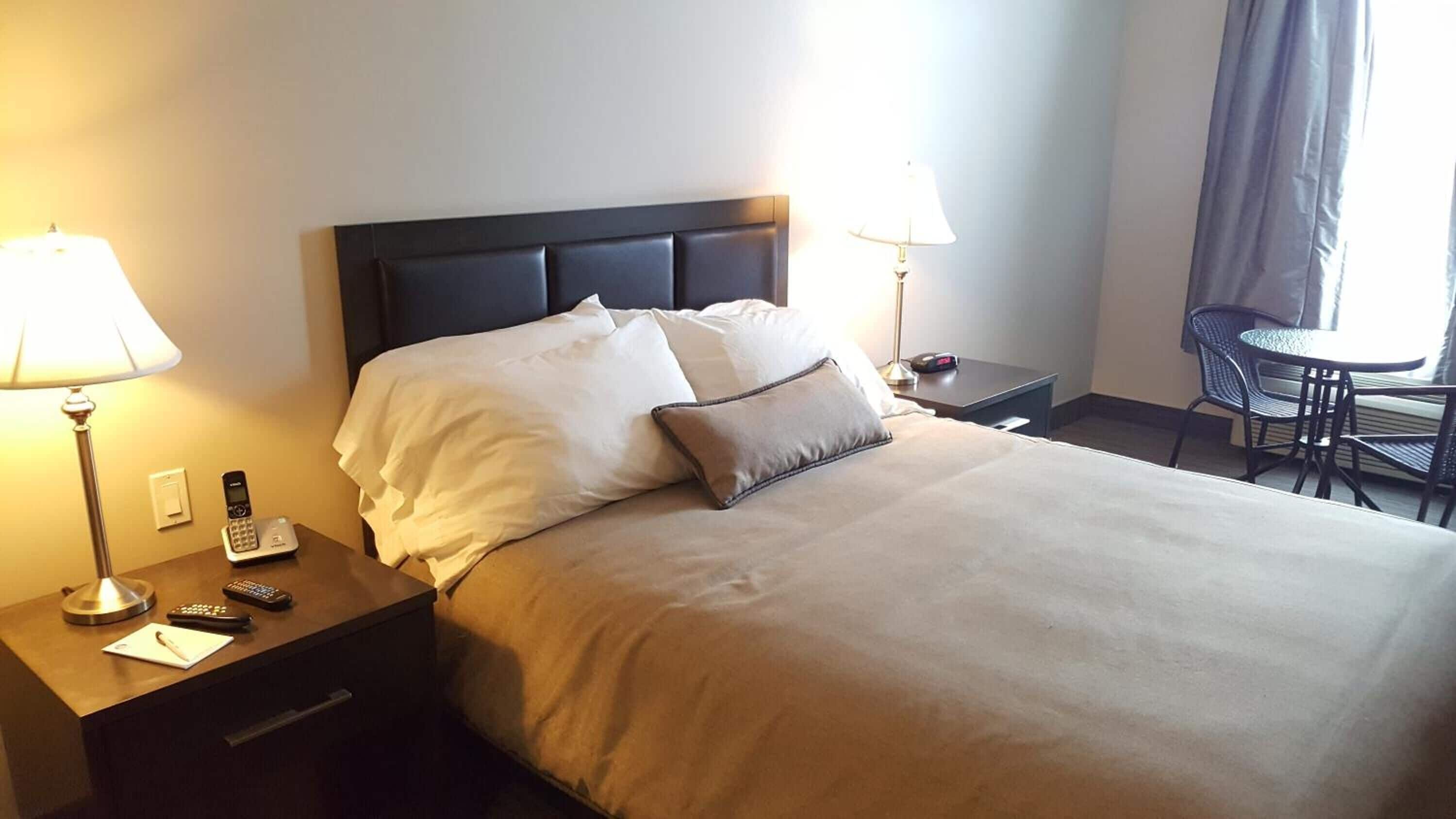Habitación de hotel con cama, lámparas de noche, escritorio con teléfono y controles remotos, y una pequeña mesa con una silla.
