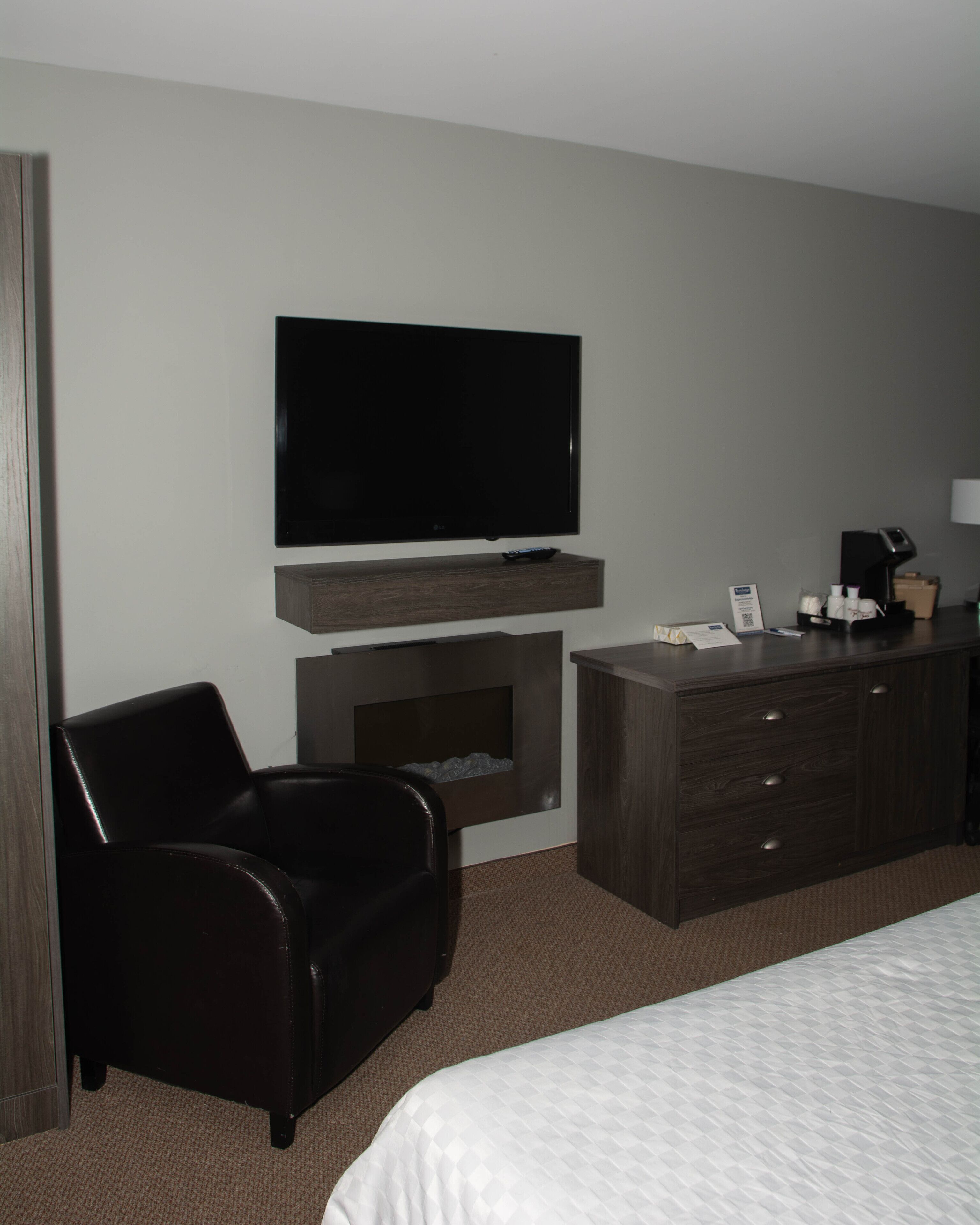 Habitación de hotel con televisor de pantalla plana sobre una consola de madera, un sillón de cuero, una cama con colcha blanca y una pequeña chimenea.
