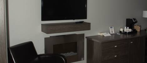 Habitación de hotel con televisor de pantalla plana sobre una consola de madera, un sillón de cuero, una cama con colcha blanca y una pequeña chimenea.