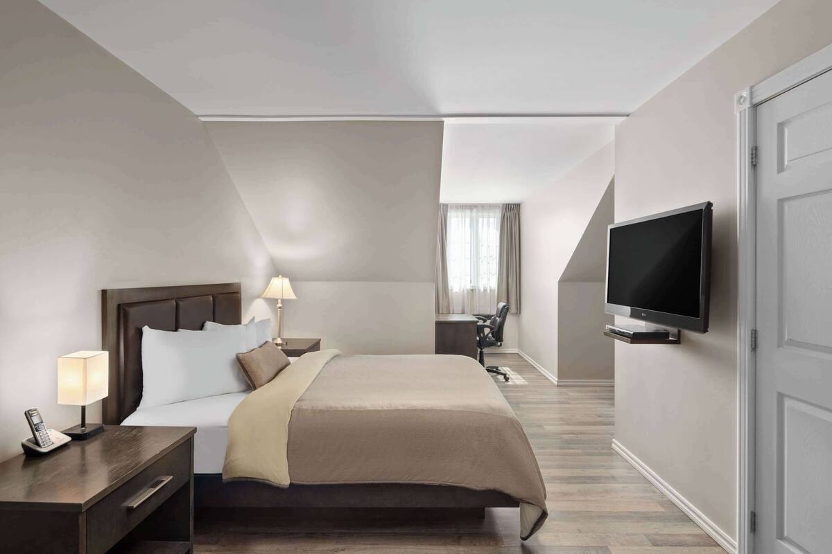 A hotel room with a bed, bedside lamps, a desk, a chair, a television, and a window with curtains.