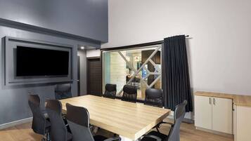 A conference room with a large table, six chairs, a flat-screen TV, and a glass partition with a view of a kitchen area.