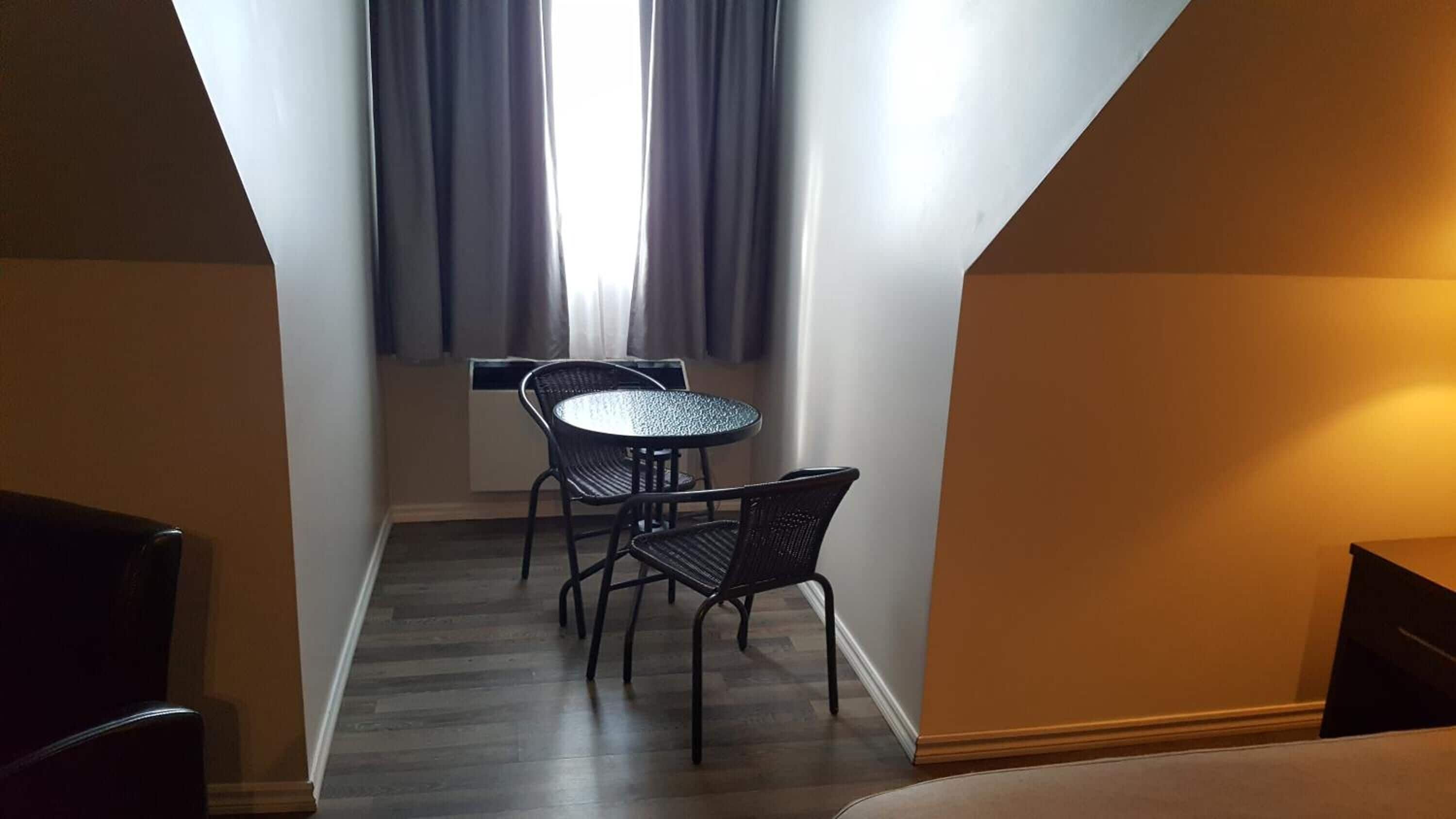 Um quarto pequeno e estreito, com uma mesa redonda e duas cadeiras, uma janela com cortinas, e teto inclinado.