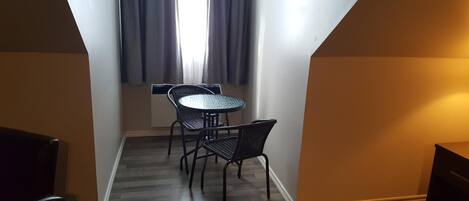 Um quarto pequeno e estreito, com uma mesa redonda e duas cadeiras, uma janela com cortinas, e teto inclinado.