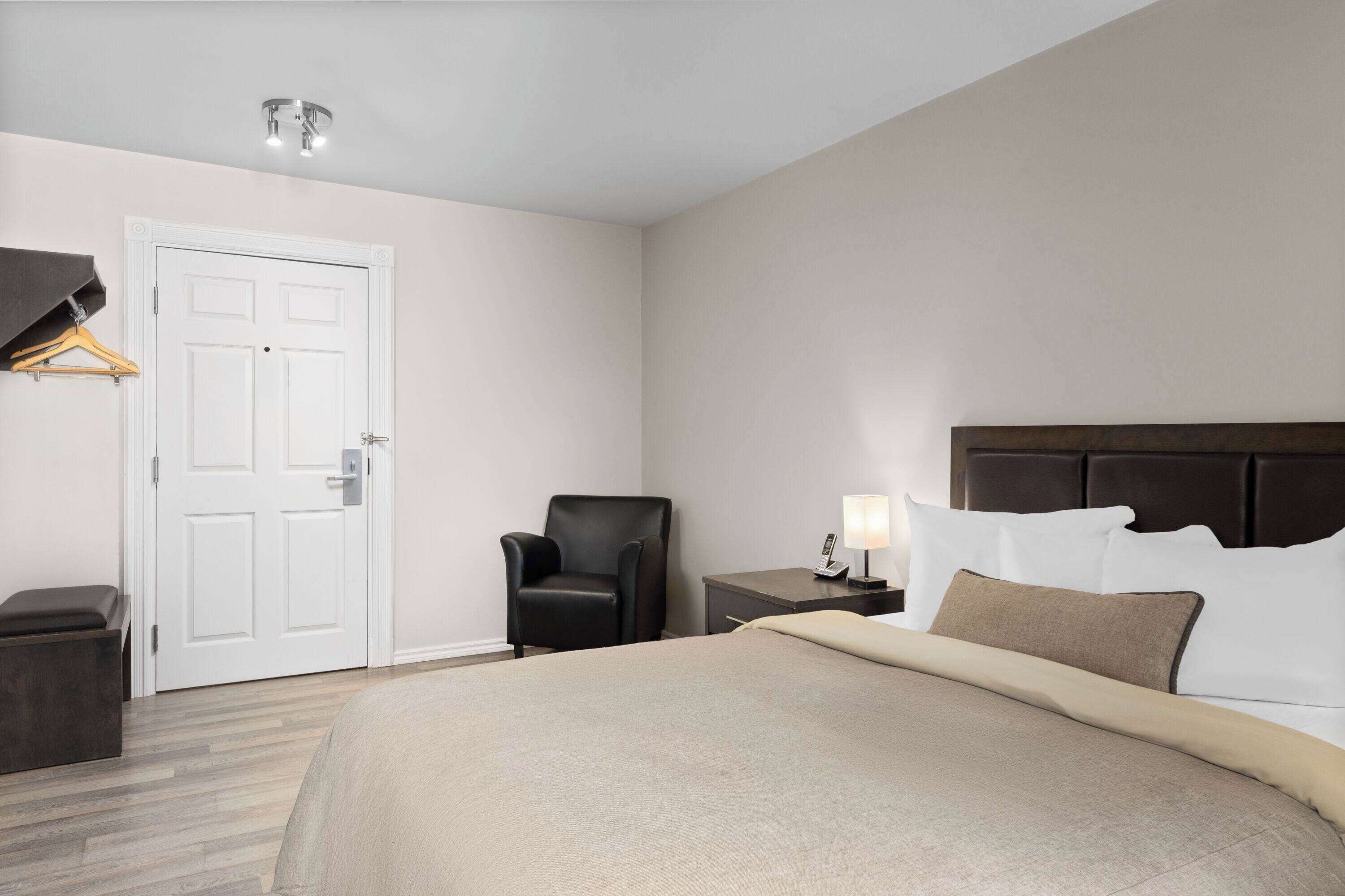 A hotel room with a bed, a chair, a desk, a lamp, and a coat rack.