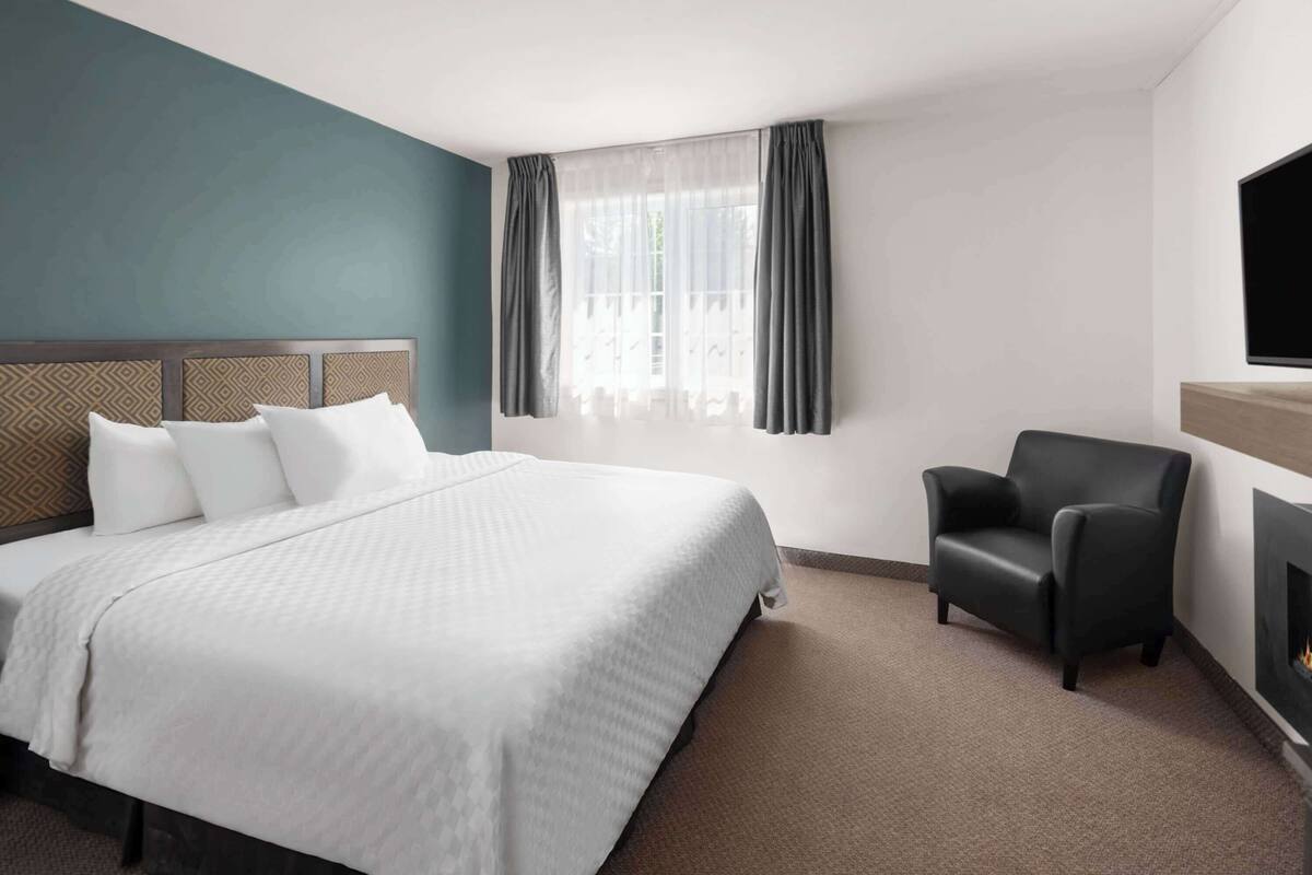 A hotel room with a large bed, a chair, a fireplace, and a window with curtains.