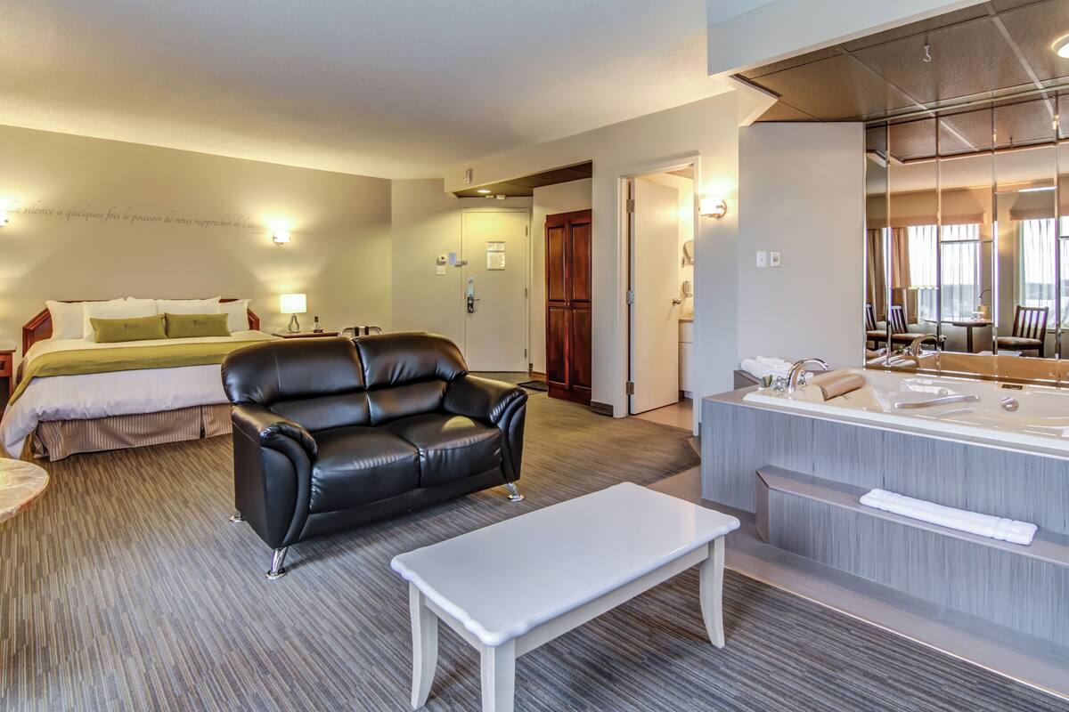 A hotel room with a bed, a black leather sofa, a white coffee table, a bathroom with a large mirror and bathtub, and a door leading to another room.