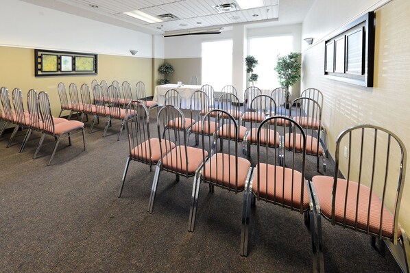 A conference room with rows of metal chairs and a table, carpeted floor, and framed pictures on the wall. - Hôtel du Jardin (St. Felicien)