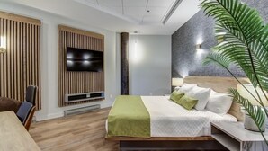 Une chambre d’hôtel moderne avec un plancher en bois, un lit recouvert d’une literie verte et blanche, un téléviseur à écran plat fixé sur un panneau en bois, et une grande plante en pot.