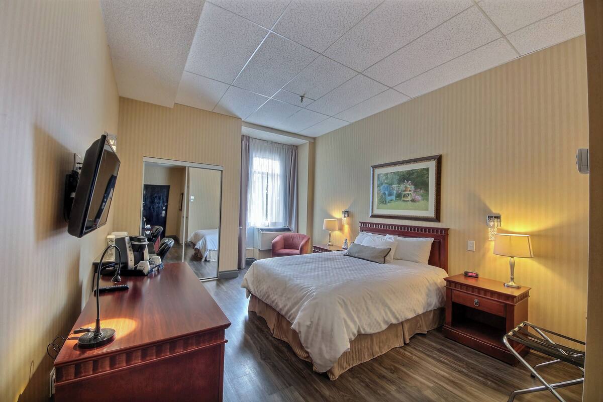 A hotel room with a bed, bedside table, lamp, and a desk with a computer and phone.