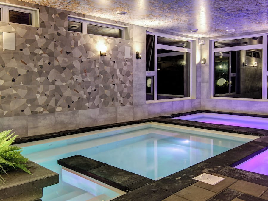 Una piscina interior moderna con azulejos geométricos en las paredes, iluminación integrada y un área de piscina transparente.