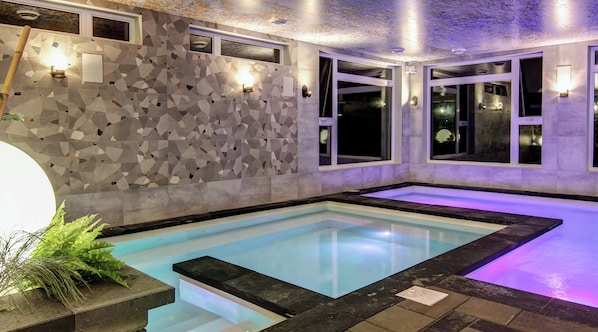 Una piscina interior moderna con azulejos geométricos en las paredes, iluminación integrada y un área de piscina transparente.