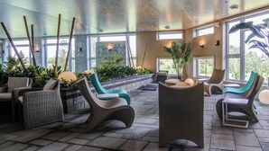 Un espace de détente intérieur moderne, doté de sièges et de plantes, de grandes fenêtres et d’un sol carrelé.