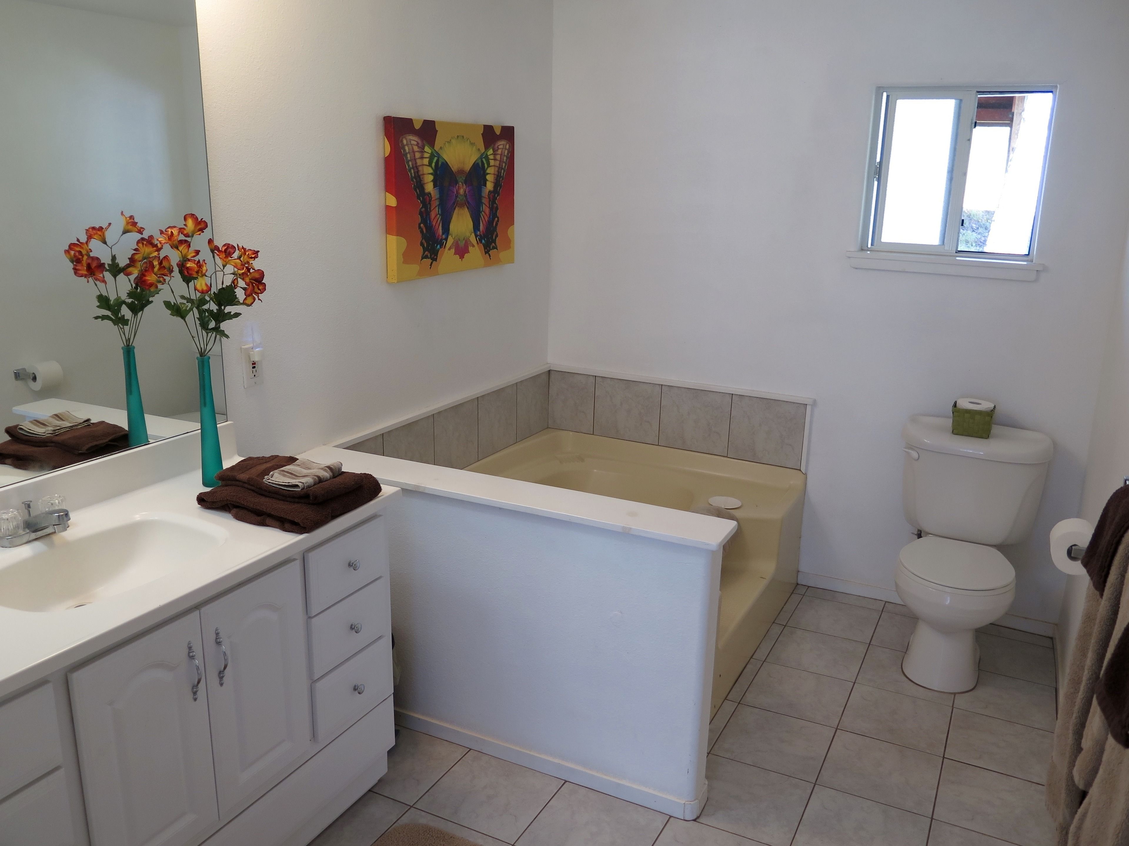 A modern bathroom with a bathtub, toilet, and sink. There is a mirror, a vase with flowers, and a colorful painting on the wall.