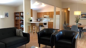 Un salón con un sofá negro, dos sillones negros, una mesa de centro de cristal y una estantería. Cuenta con una zona de cocina con armarios de madera, un refrigerador y una mesa de comedor con sillas.
