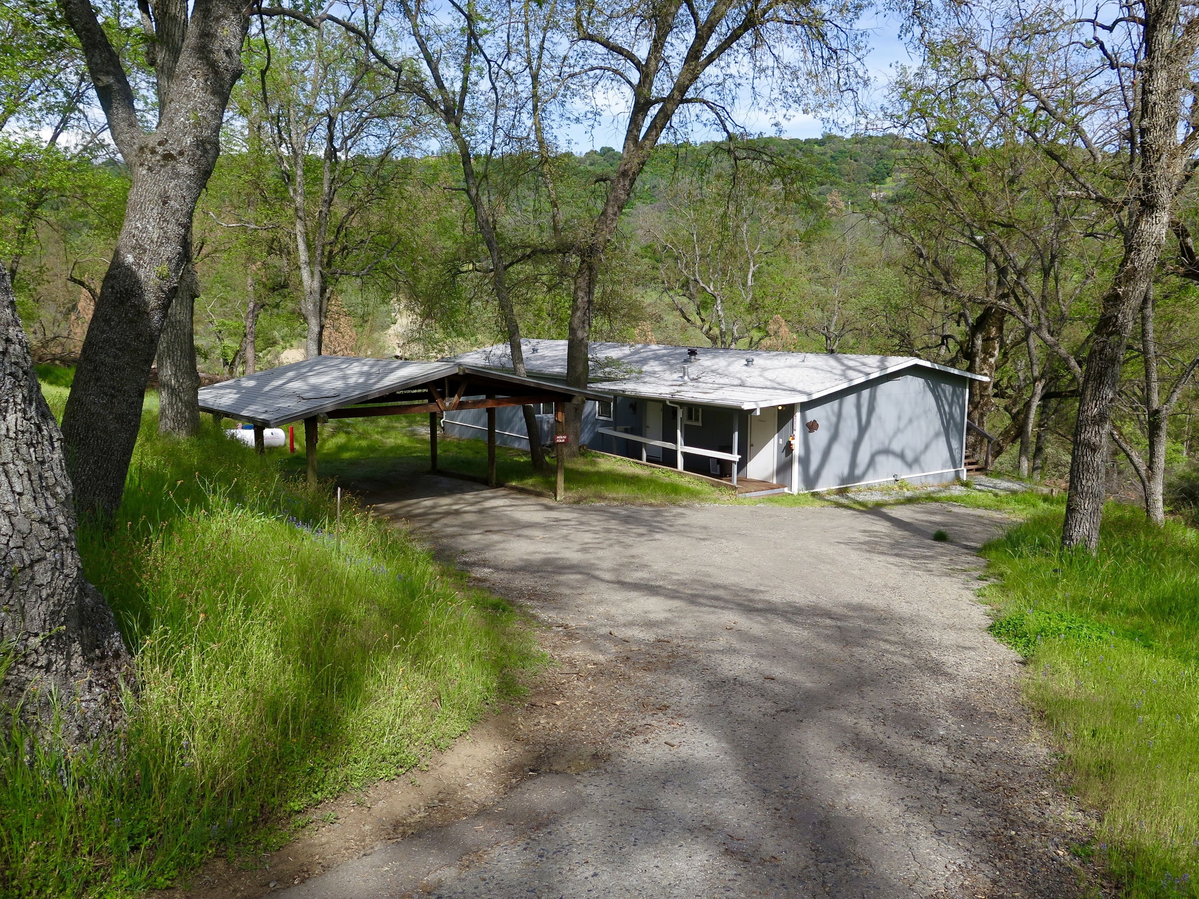 A rustic cabin with a covered porch and a separate garage, surrounded by dense trees and a dirt path.