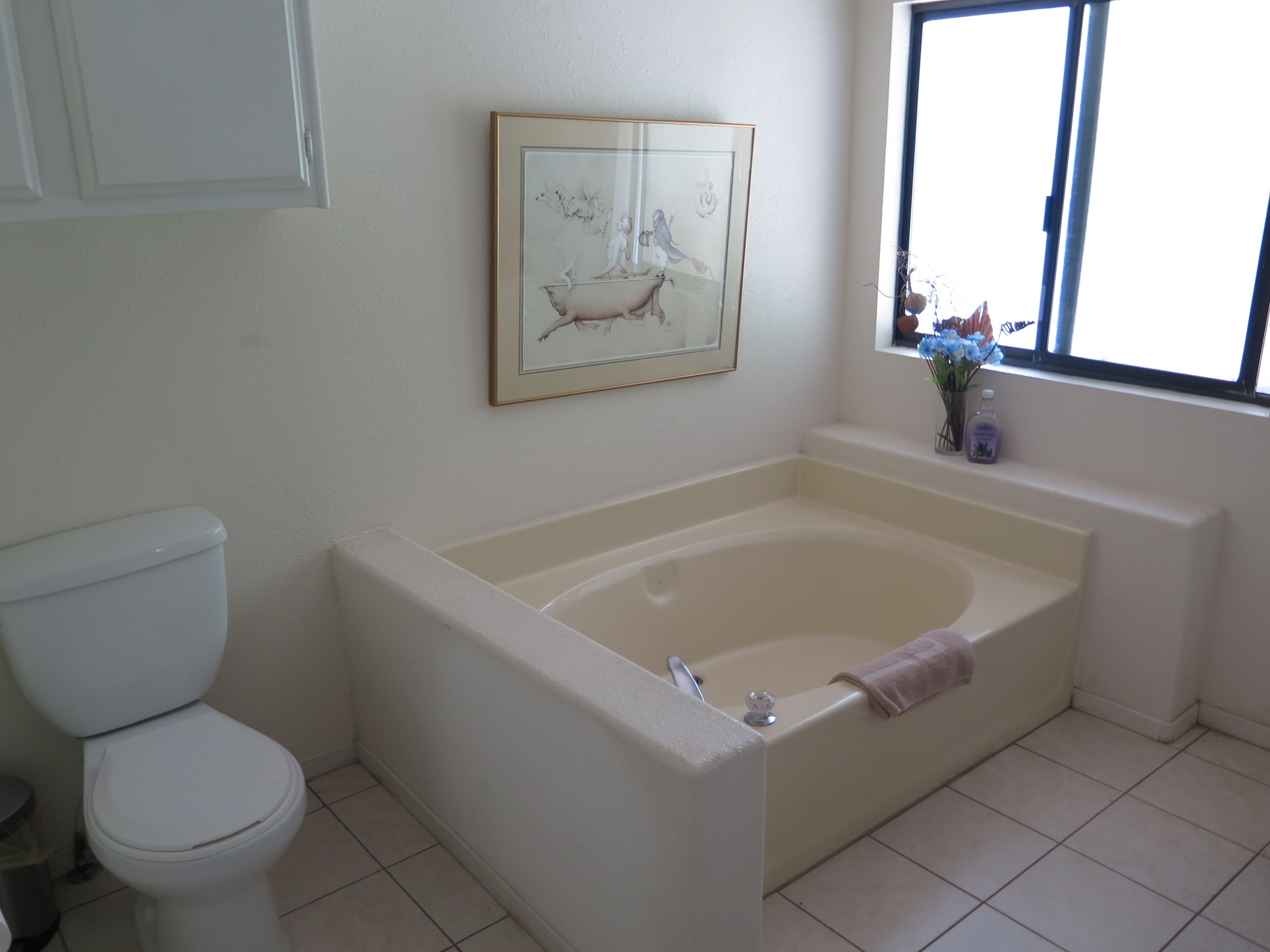 A bathroom with a toilet, a bathtub, a framed artwork on the wall, and a window with flowers.