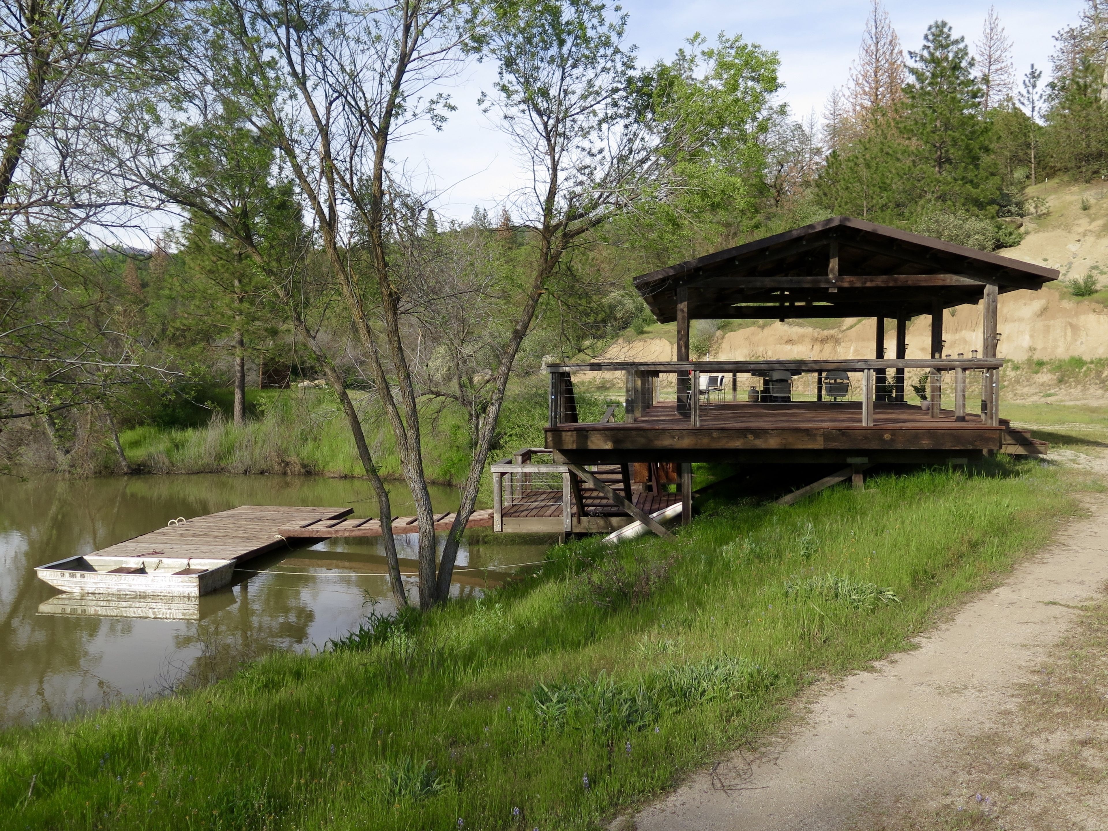 Une terrasse en bois avec un espace salon couvert donnant sur une rivière paisible, un quai avec un petit bateau, et un sentier de terre qui longe la rivière.