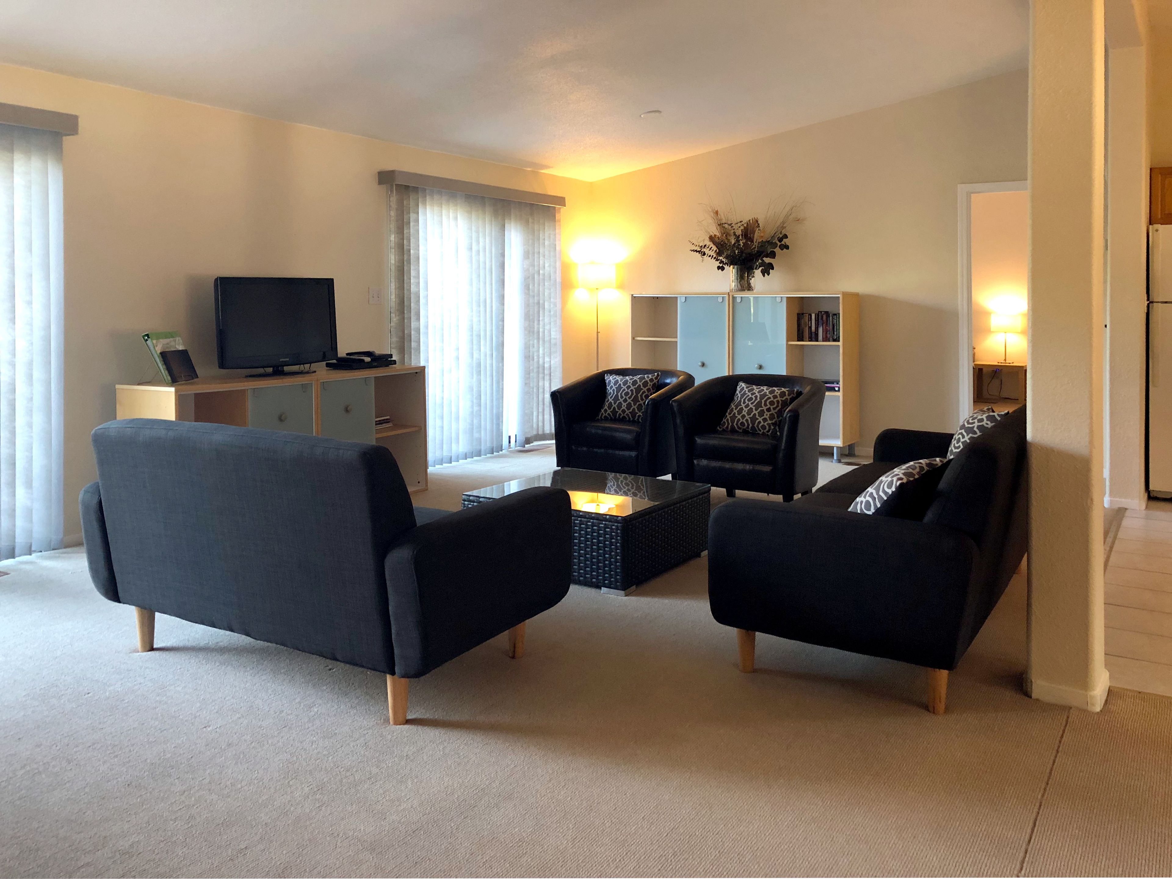 A modern living room with a flat-screen TV, two black sofas, a coffee table, and a bookshelf.