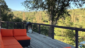 Una terraza de madera con cojines anaranjados y una parrilla, con vistas a un área boscosa.