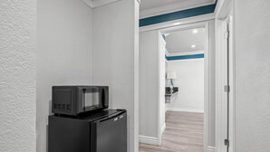 A compact kitchen with a black refrigerator and microwave, a wooden floor, and a hallway leading to a room with a blue accent wall and a lamp. - Rodeway Inn Commerce - Los Angeles (Los Angeles)