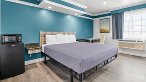 A compact hotel room with teal walls, a bed with white bedding, a microwave, a small table, a chair, and an air conditioning unit. - Rodeway Inn Commerce - Los Angeles (Los Angeles)