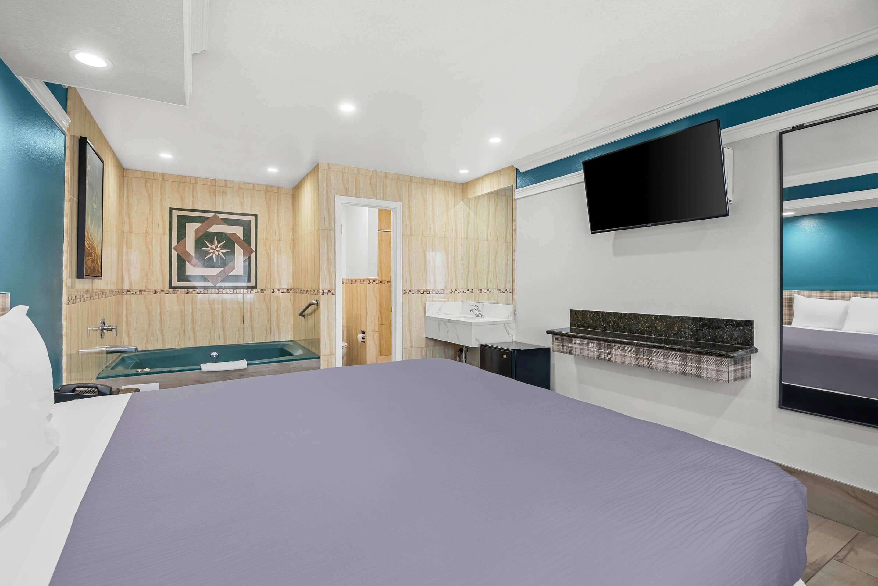A modern hotel room with a large bed, a built-in TV, a bathroom with a bathtub, and a small desk.