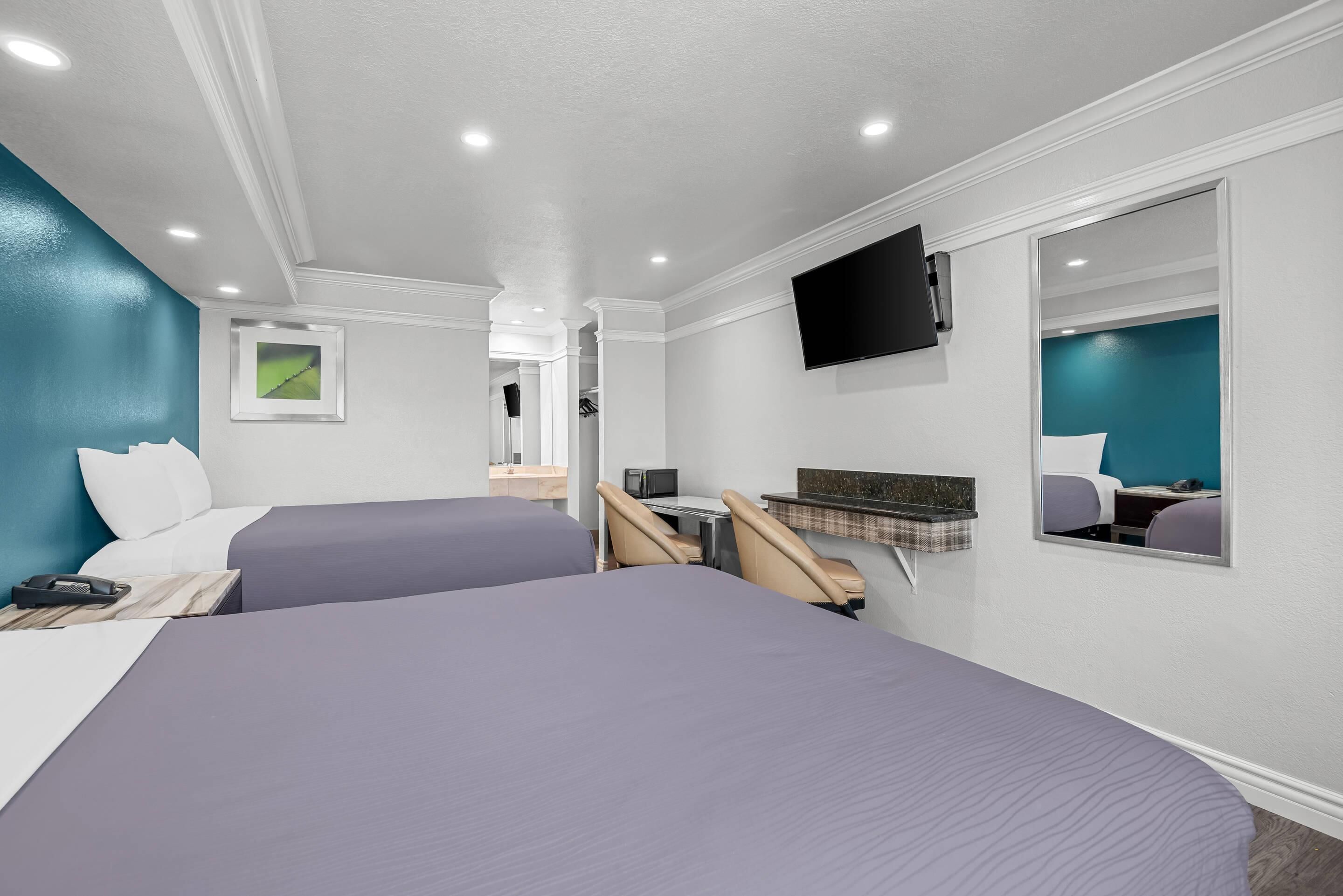 A hotel room with two beds, a desk with a chair, a flat-screen TV mounted on the wall, and a small seating area with a bench and cushions.
