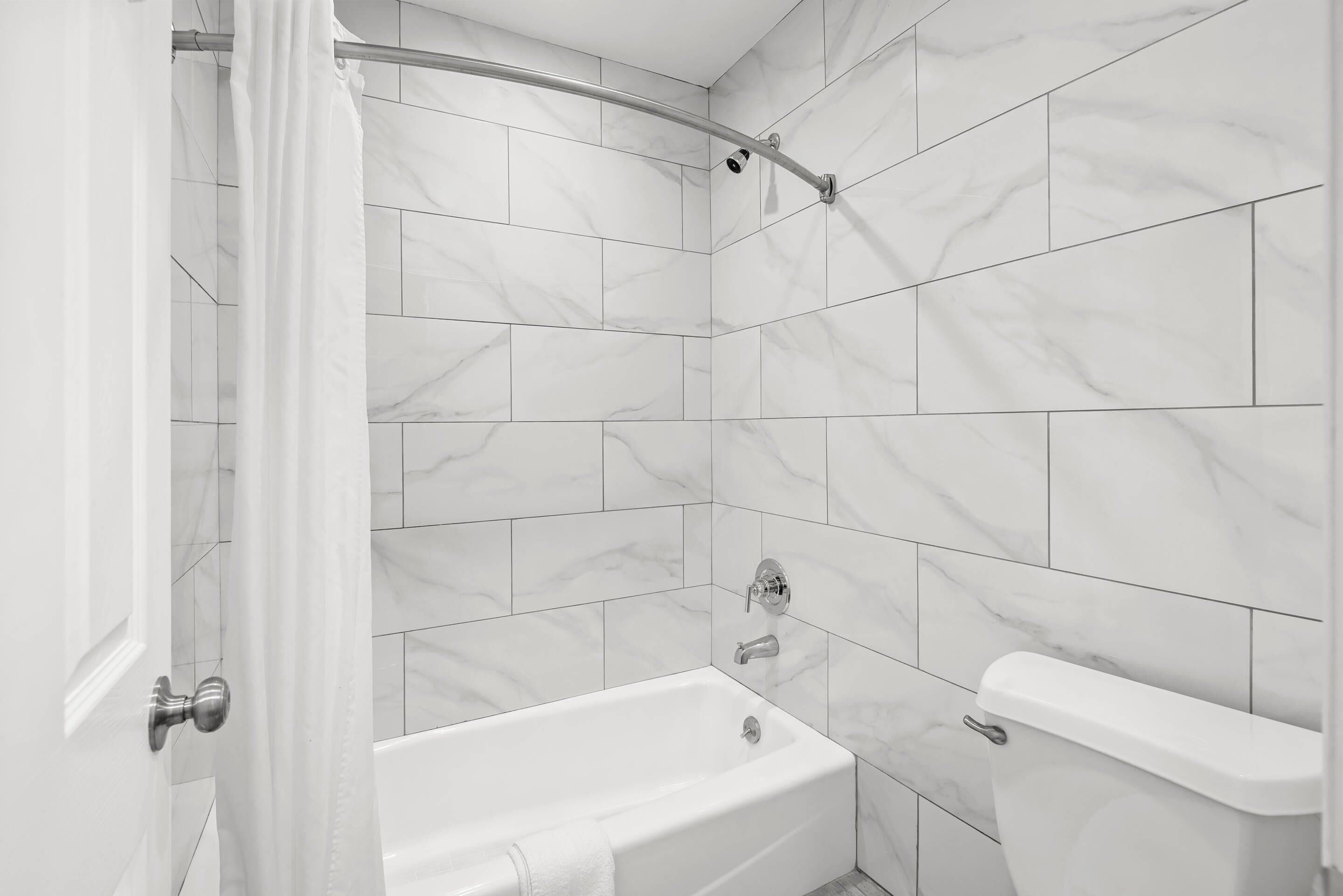 Baño con bañera, inodoro y zona de ducha, con azulejos de mármol blanco y cabezal de ducha curvo.