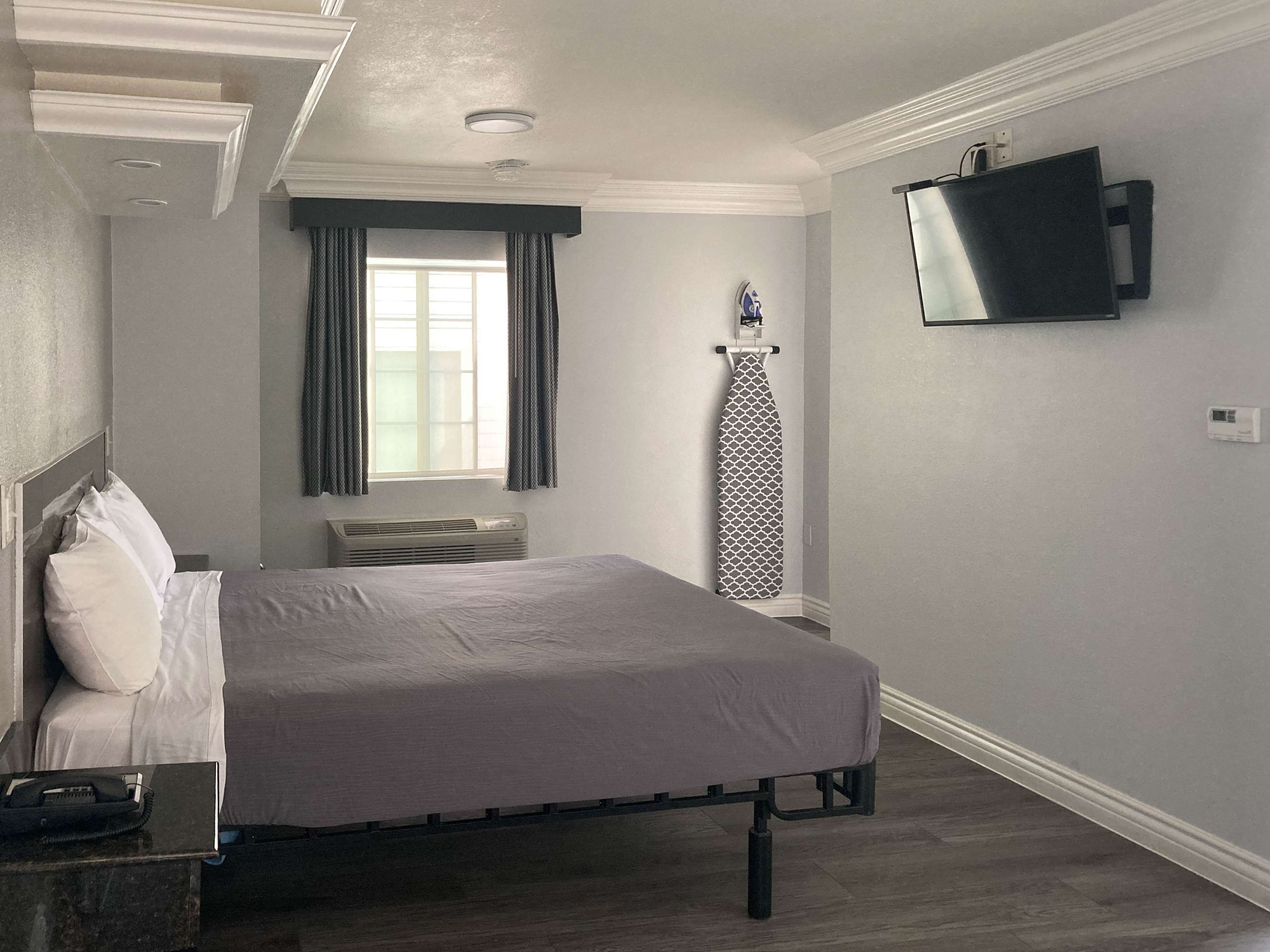 A bedroom with a bed, a flat-screen TV mounted on the wall, a window with curtains, and an iron stand.