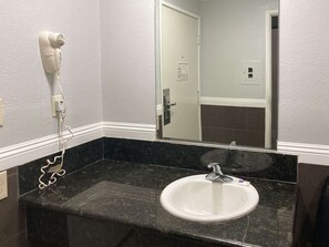 A bathroom with a white sink, a mirror, and a hair dryer plugged into the wall. - Motel 6 Glendale, CA – Pasadena Burbank Los Angeles (Glendale)