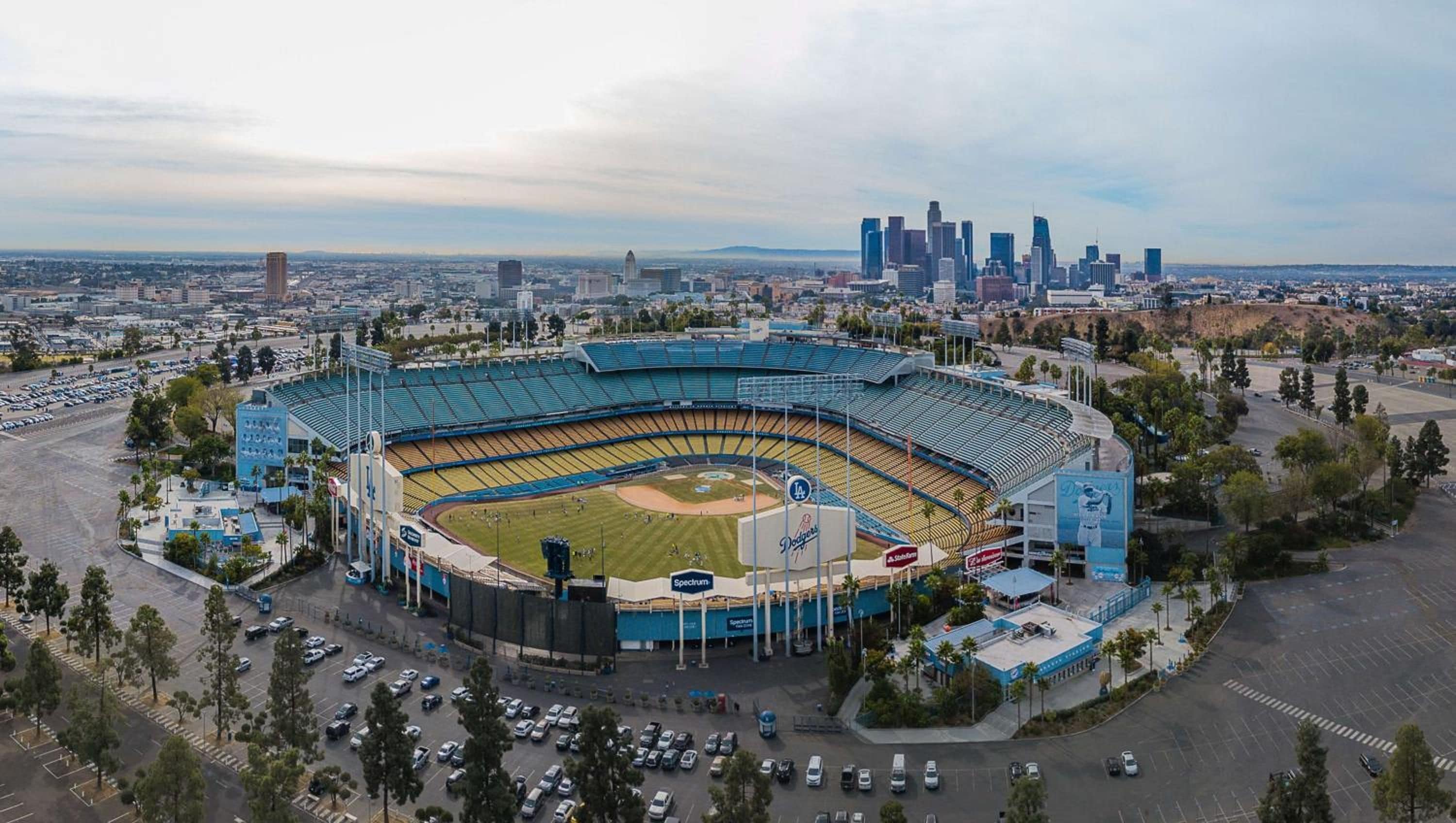 A large stadium with a baseball field, surrounded by parking areas and a city skyline in the background.