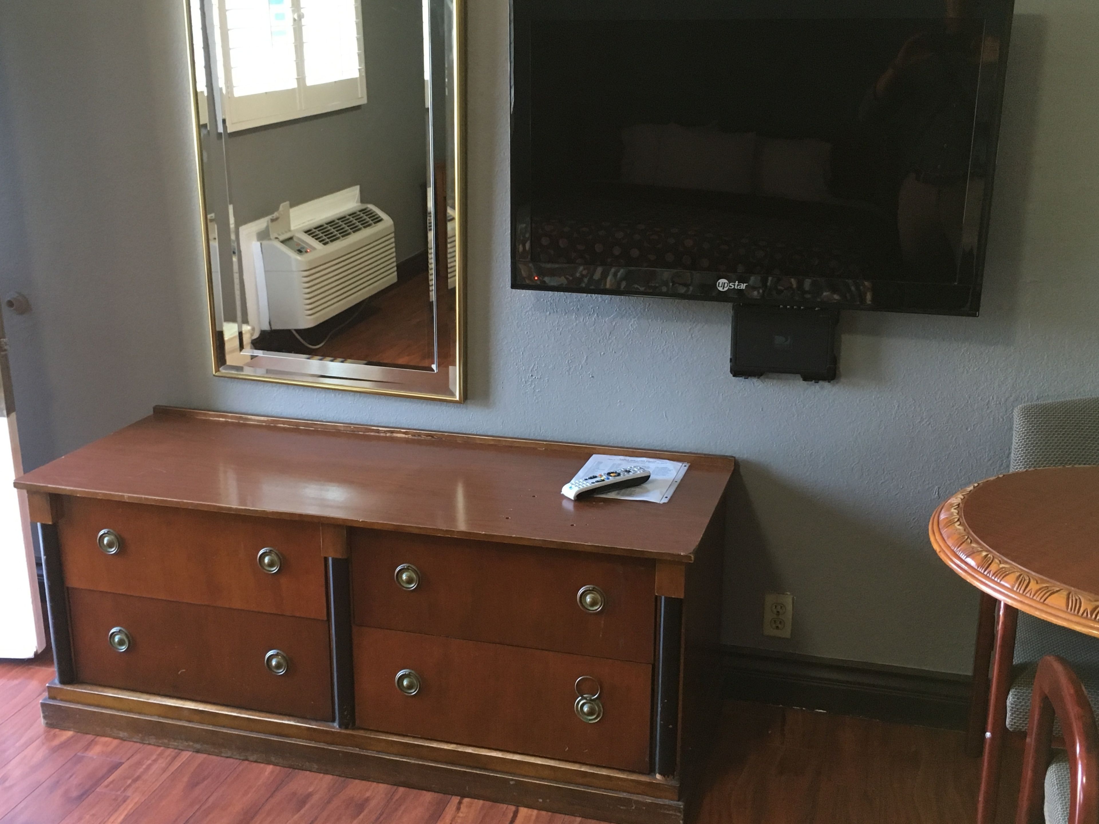 Una habitación con un aparador de madera, un espejo, un televisor montado en la pared y una mesa redonda.