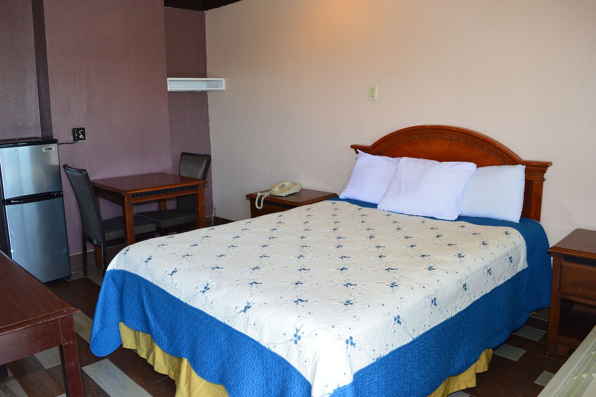 A hotel room with a bed, a desk, a chair, a refrigerator, and a telephone.