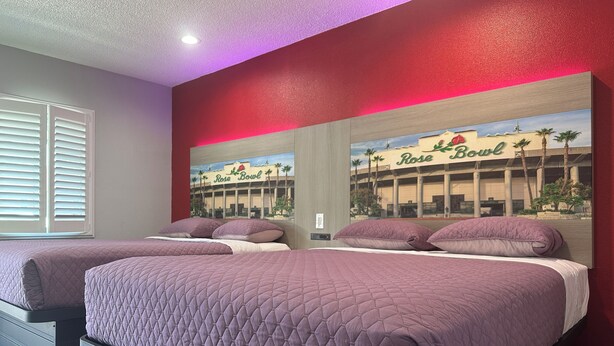 A hotel room with two beds, a large window with blinds, and a mural of a building with palm trees. - Hi-Way Host Motel (Pasadena)
