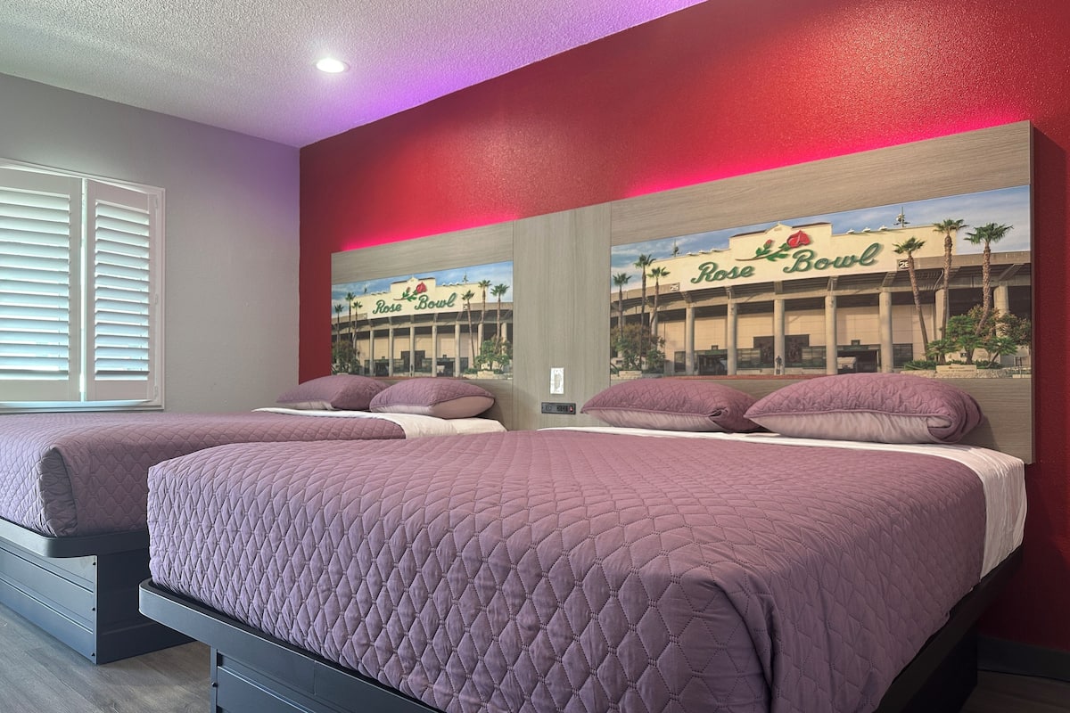 A hotel room with two beds, a large window with blinds, and a mural of a building with palm trees.