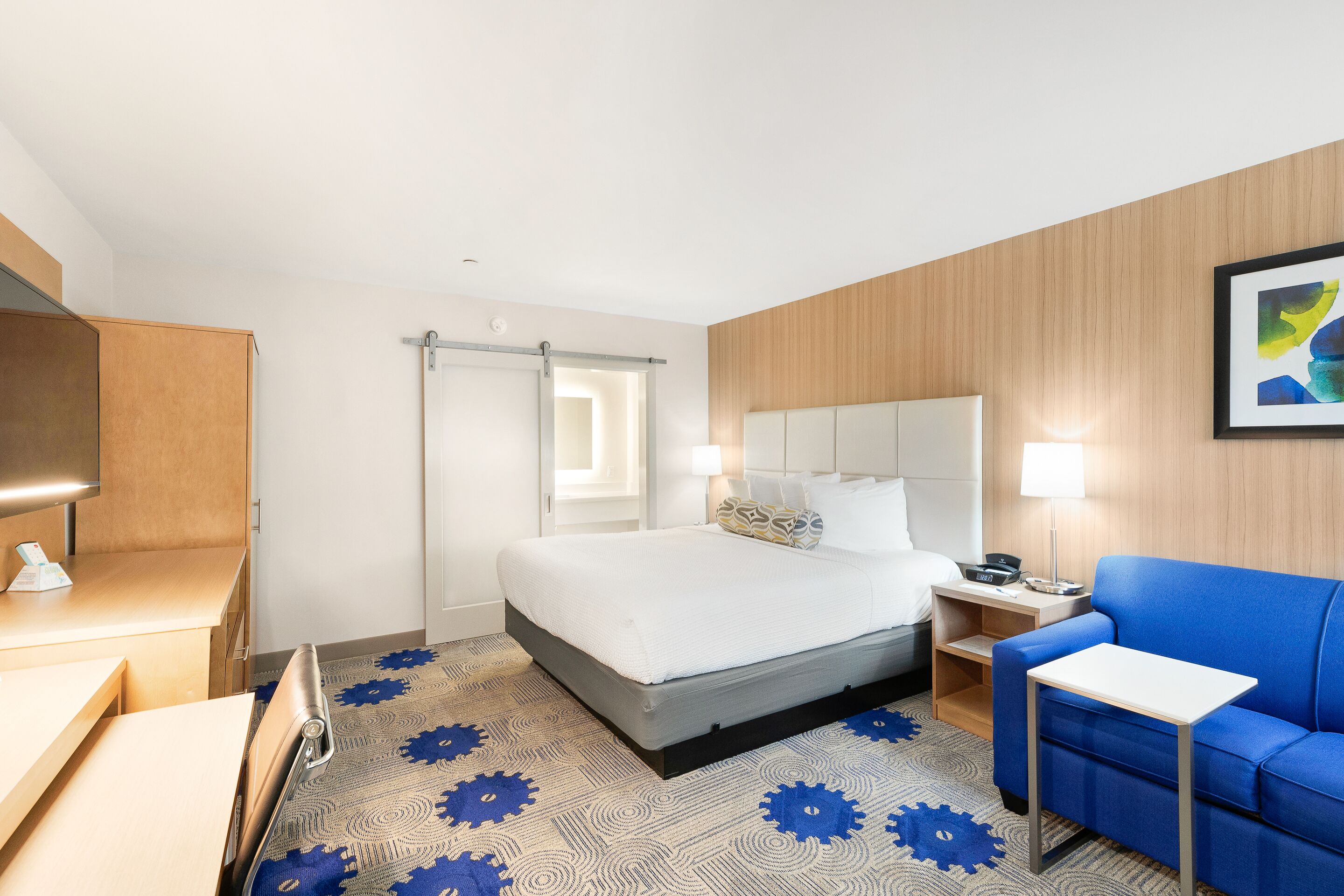 A hotel room with a bed, a blue sofa, a small table, a nightstand, and a wall-mounted TV.
