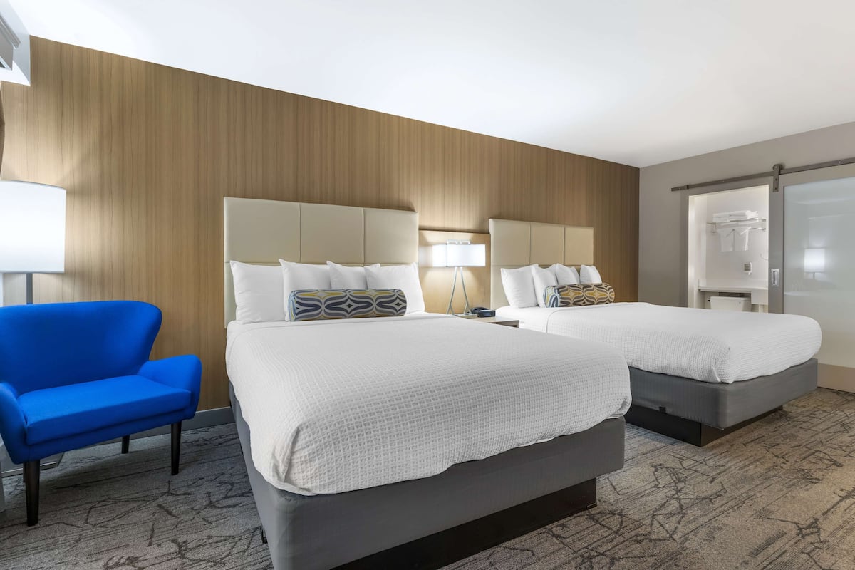 Une chambre d’hôtel avec deux lits, un fauteuil bleu et un grand miroir.