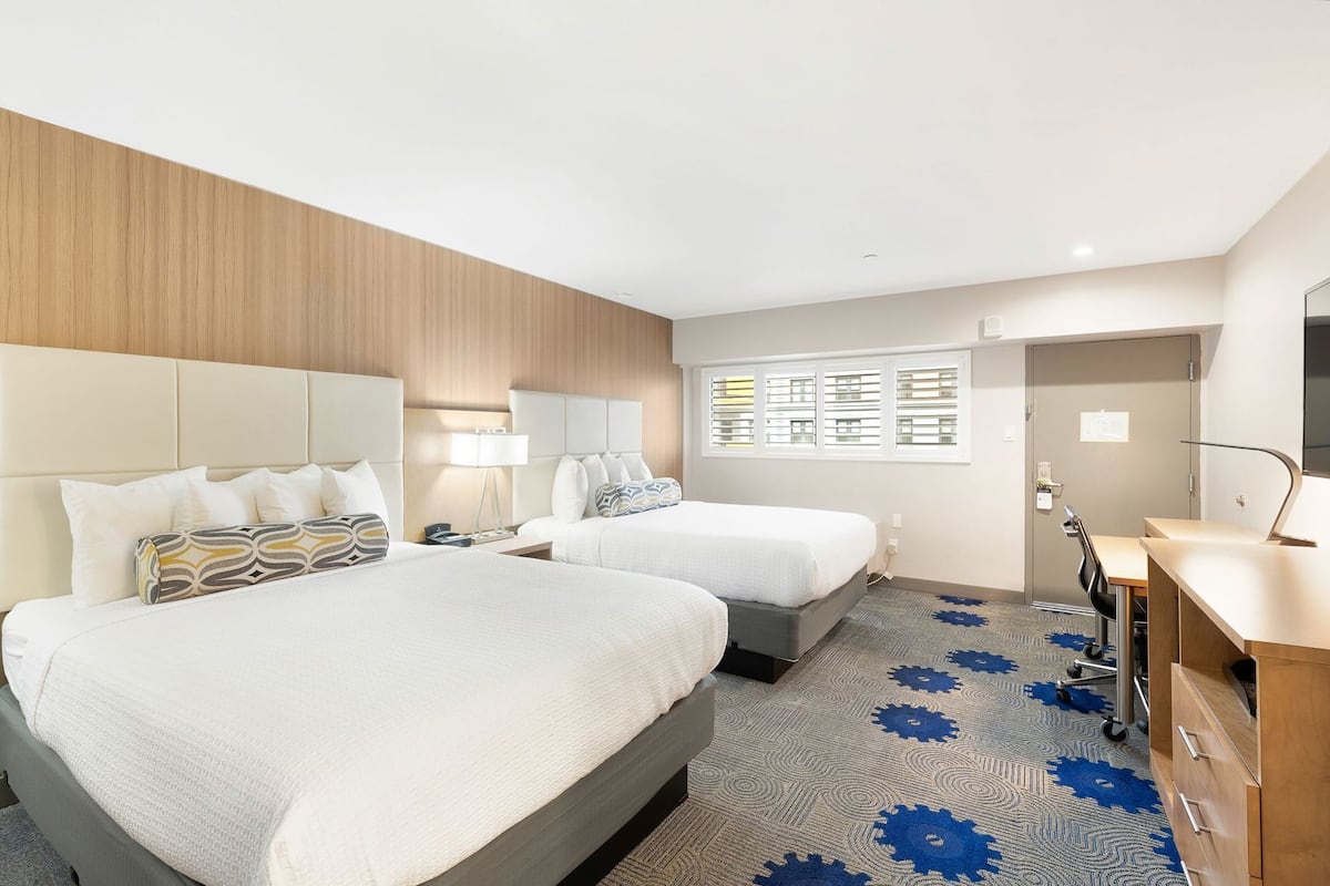 Une chambre d’hôtel avec deux lits, un bureau, une chaise, une lampe, une fenêtre avec des stores et une porte.