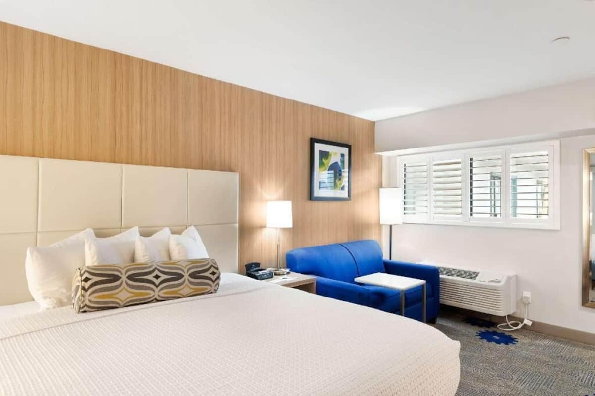 Une chambre d’hôtel avec un grand lit, un fauteuil bleu, une petite table, une lampe et une fenêtre avec des stores.