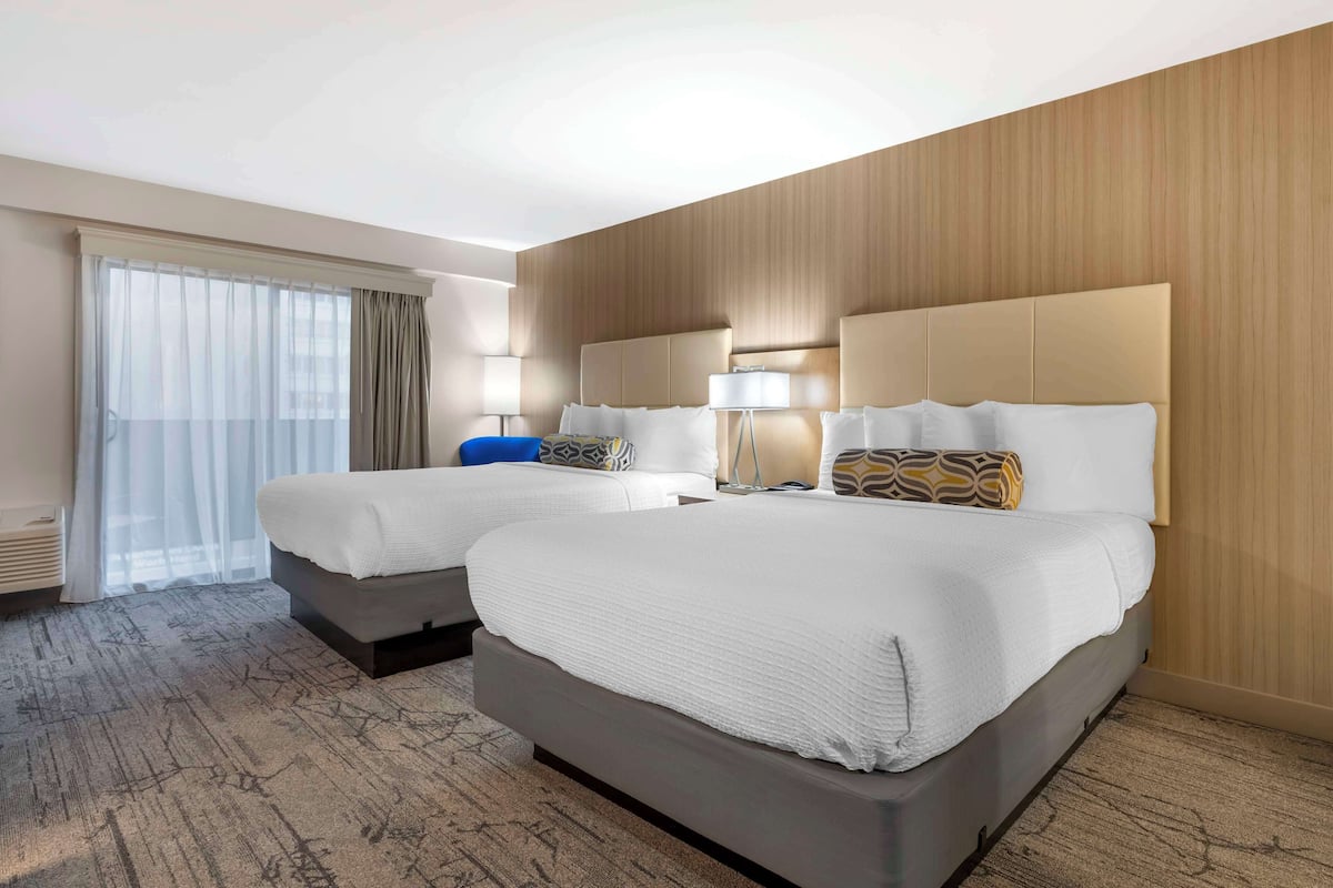 Une chambre d’hôtel avec deux lits, une grande fenêtre avec des rideaux légers, un mur en bois et un sol recouvert de moquette.