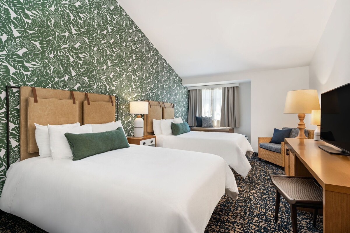 A hotel room with two beds, a desk with a TV, a window with curtains, and a patterned wall.