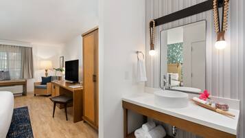 A hotel room with a bed, a desk, a chair, a TV, a large mirror, a sink, and a window with curtains.