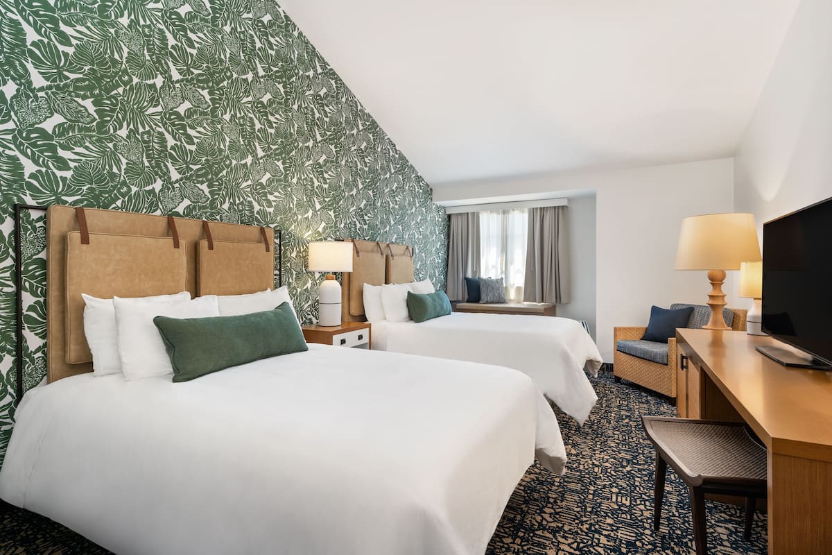 A hotel room with two beds, a desk, a TV, and a window with curtains.