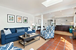 Uma sala de estar moderna com um sofá azul, poltrona estampada e uma mesa de centro. Uma área de cozinha é visível ao fundo, com uma placa que diz "Mason Beach Inn".