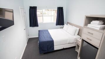 A hotel room with a bed, a TV mounted on the wall, a small desk with a drawer, a mini fridge, and a window with curtains.
