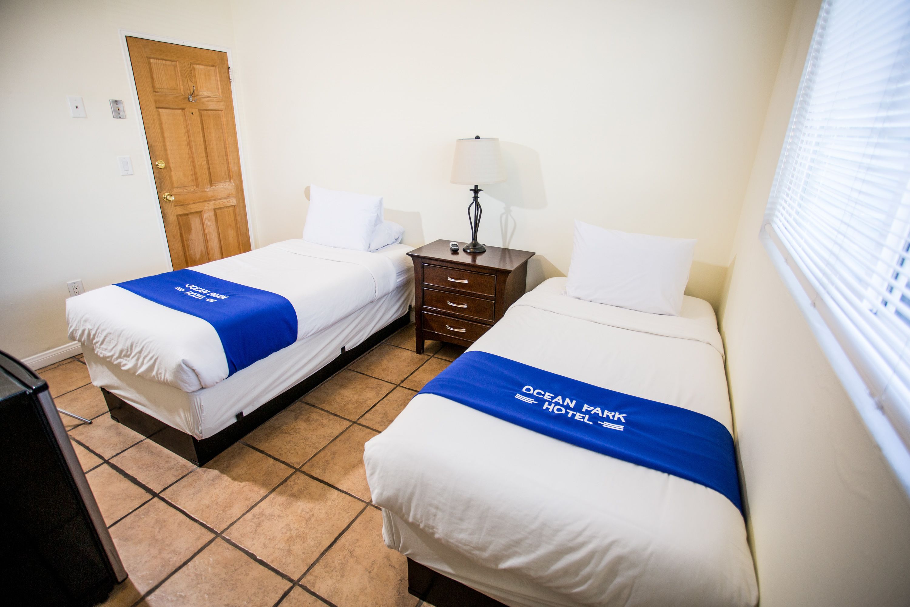 Habitación de hotel con dos camas, una puerta de madera, una mesita de noche con lámpara y una ventana con persianas.
