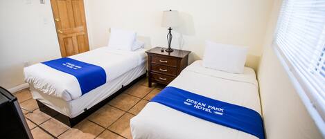 Habitación de hotel con dos camas, una puerta de madera, una mesita de noche con lámpara y una ventana con persianas.