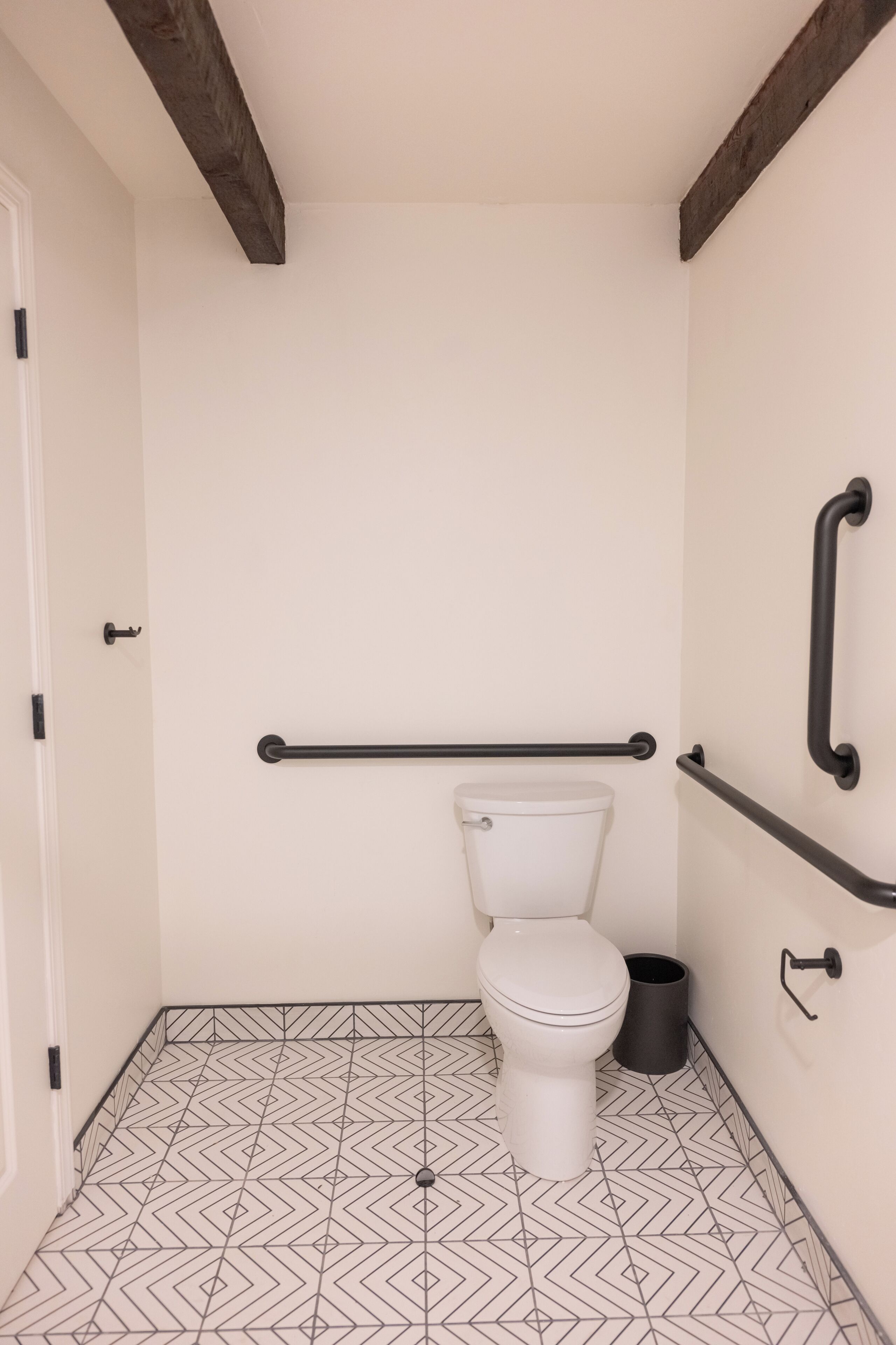 A small, modern bathroom with a white toilet, black grab bars, and a geometric patterned floor.