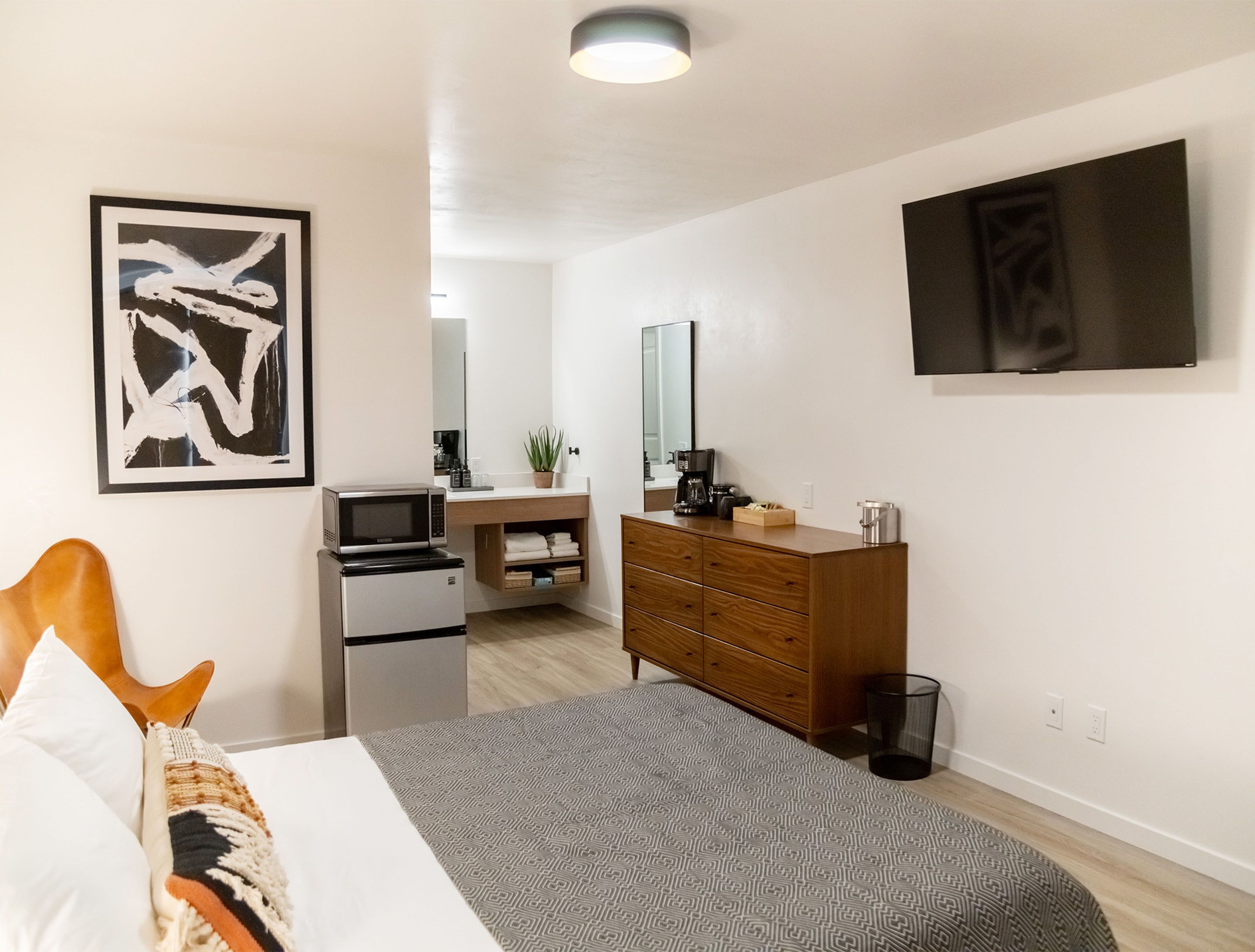 A modern bedroom with a bed, a dresser, a microwave, a coffee maker, and a large mirror.