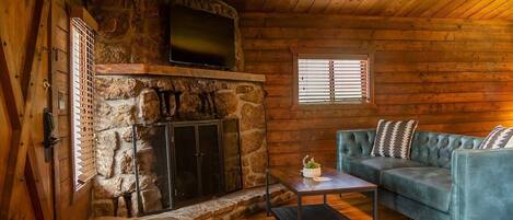 Interior de una cabaña de troncos con chimenea de piedra, un televisor montado encima y un sofá con cojines estampados, junto a una pequeña mesa con una planta.